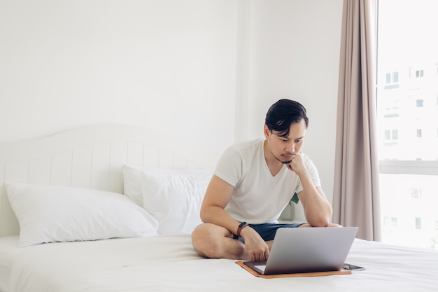El hombre está sentado en la cama y trabaja en su computadora portátil.