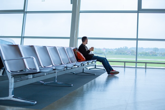 Un hombre está sentado en una fila de asientos vacíos frente a una gran vidriera en una terminal del aeropuerto, esperando un vuelo.