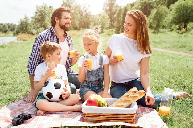 El hombre está sentado sobre una manta y mirando a su esposa. Ella lo está mirando. Los niños están sonriendo el uno al otro. Todos tienen un vaso de jugo.