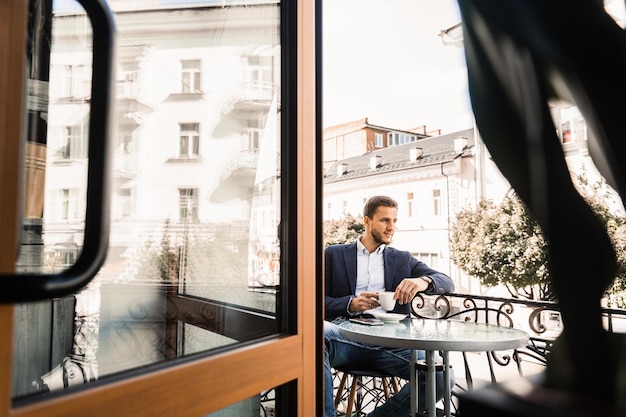 El hombre está sentado en la terraza de verano en la cafetería tomando café y sonriendo Guapo con café