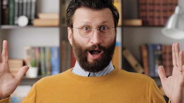 El hombre está sorprendido, consternado. Sonriente hombre barbudo con gafas en la habitación de la oficina o apartamento mirando a cámara y agita sus manos emocionalmente expresando su increíble sorpresa. Vista cercana