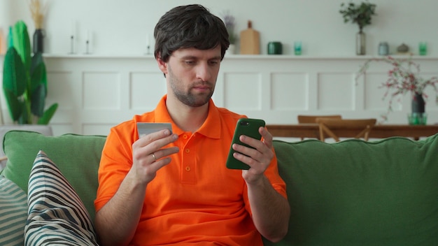 El hombre está sosteniendo una tarjeta de crédito y está usando un teléfono inteligente para hacer compras en línea en casa