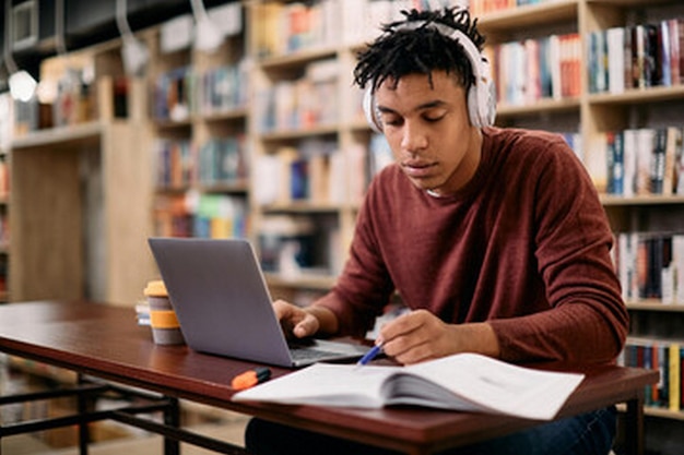 un hombre está trabajando en una computadora portátil en una biblioteca con un libro en su regazo