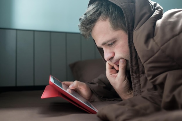 El hombre está trabajando de noche acostado en el sofá de la sala de estar con una tableta que cubre su cabeza con una manta