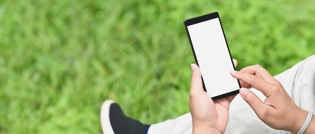 Un hombre está usando un teléfono inteligente de pantalla en blanco mientras está sentado sobre el campo de hierba como fondo.