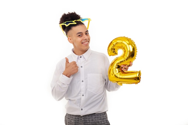 Un hombre estadounidense feliz con gafas de estrella en la cabeza mostrando los pulgares hacia arriba sosteniendo un globo con forma de número dos