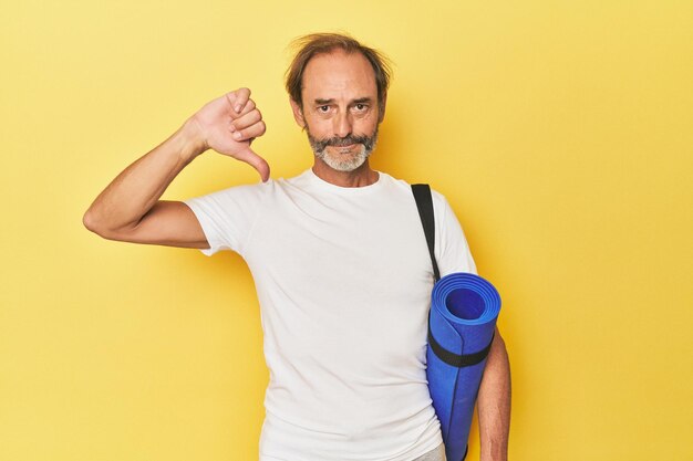 Hombre con estera de yoga en estudio amarillo mostrando un gesto de desagrado pulgares hacia abajo Concepto de desacuerdo