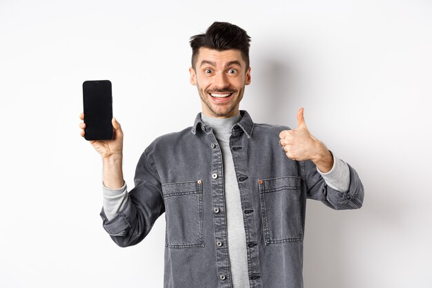 Hombre con estilo alegre que muestra la pantalla vacía del teléfono inteligente y el pulgar hacia arriba, el concepto de compras en línea y comercio electrónico.