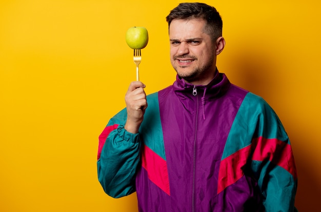 Hombre de estilo en chándal de los 90 con manzana en horquilla en pared amarilla