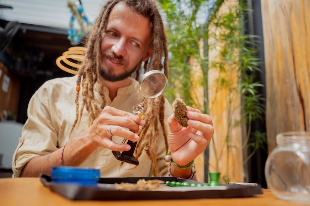 Hombre de estilo hippie examina bajo una lupa las articulaciones y cogollos de marihuana medicinal