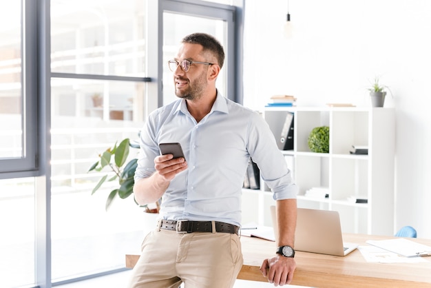 Hombre con estilo horizontal con gafas sentado en la mesa en la oficina y mirando por una ventana grande, mientras usa el teléfono móvil