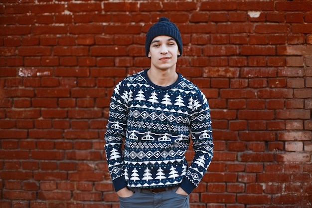 Hombre con estilo joven atractivo estadounidense en un suéter azul de punto con un adorno de Navidad en un sombrero de punto azul de invierno en jeans posando al aire libre cerca de una pared de ladrillo vintage. Chico guapo.