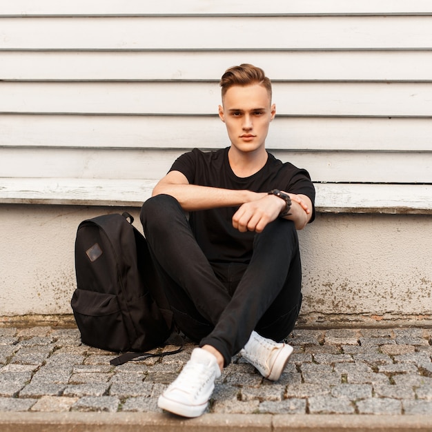 Hombre con estilo joven en camisa negra y zapatos blancos con una mochila se sienta cerca de la pared de madera
