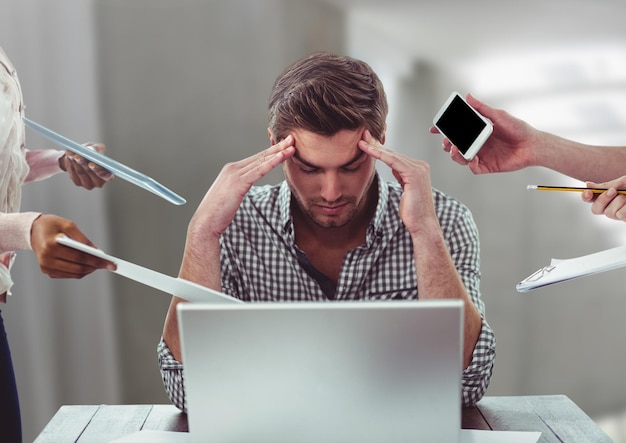 Hombre estresado en el escritorio de la computadora portátil rodeado de archivos, teléfonos, tabletas en la oficina