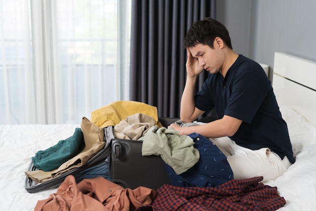 Hombre estresado que tiene problemas para empacar ropa en una maleta en el concepto de viaje de vacaciones en la cama