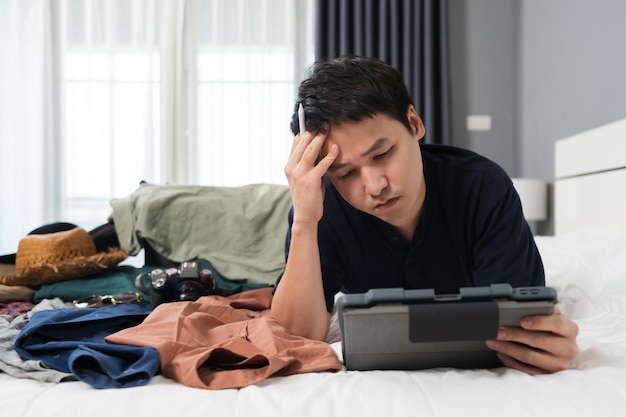 Foto hombre estresado usando tableta y empacando ropa en una maleta en un concepto de viaje de vacaciones en cama