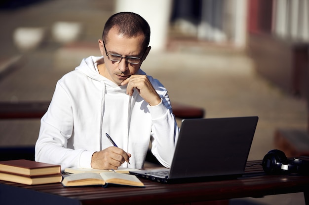 El hombre estudia en la computadora portátil y escribe en un cuaderno mientras está sentado en la calle en una mesa