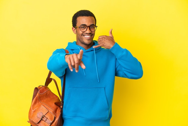 Hombre estudiante afroamericano sobre fondo amarillo aislado haciendo gesto de teléfono y apuntando hacia el frente