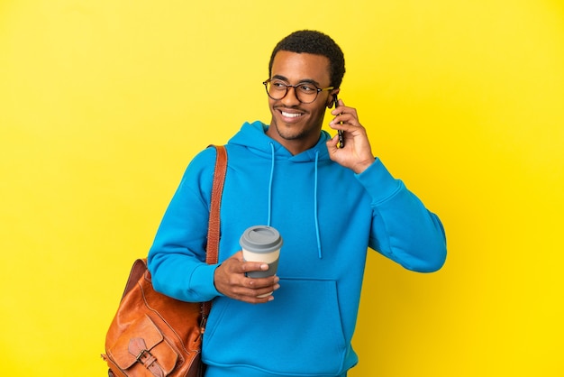 Hombre estudiante afroamericano sobre fondo amarillo aislado sosteniendo café para llevar y un móvil