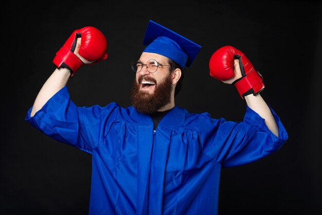 Hombre estudiante barbudo emocionado en soltero azul celebrando con guantes de boxeo