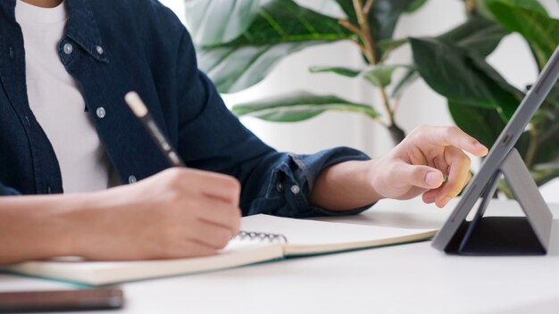 Hombre estudiante escribiendo a mano en un cuaderno mientras usa una tableta digital para e-learning