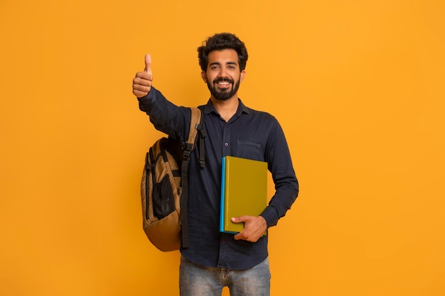 Hombre estudiante indio feliz con mochila y cuadernos mostrando pulgares hacia arriba gesto