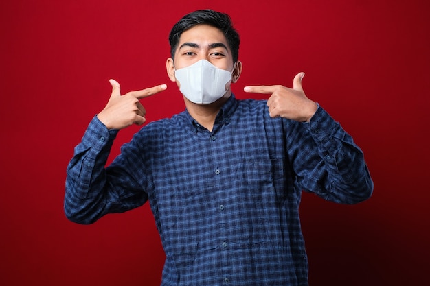 Hombre estudiante universitario asiático con el pulgar hacia arriba y máscaras para prevenir la propagación del virus Corona contra el fondo rojo mientras regresa a la escuela.