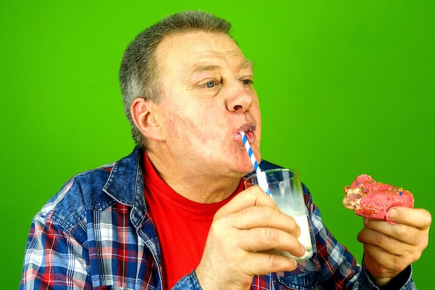 Hombre de etnia caucásica adulto mayor saboreando con placer come pastel con relleno de crema y bebe con leche de placer de vidrio transparente con tubo de cóctel. Fondo verde. De cerca.