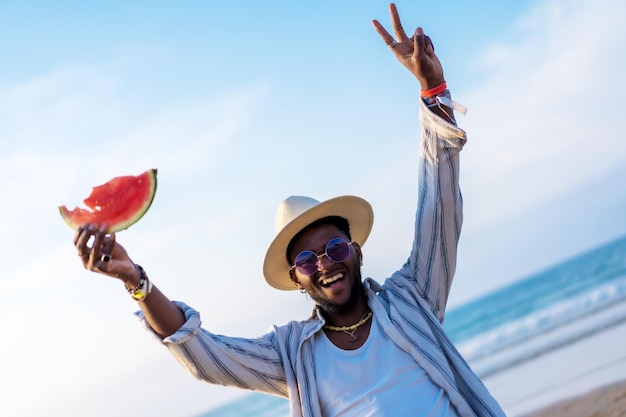 El hombre de etnia negra disfruta de las vacaciones de verano en la playa comiendo una sandía divirtiéndose