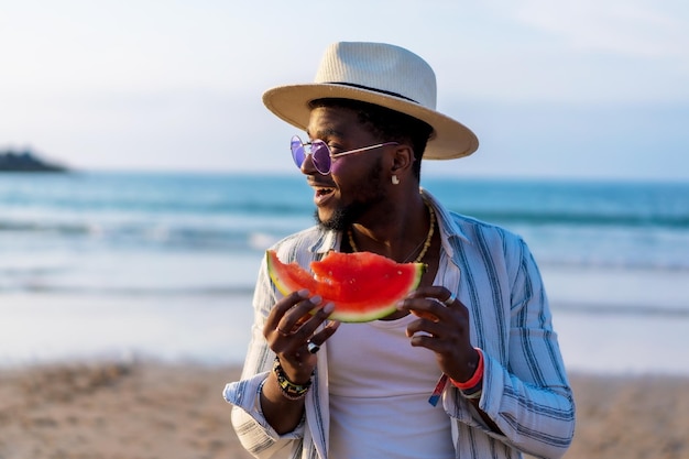 El hombre de etnia negra disfruta de las vacaciones de verano en la playa comiendo una sandía junto al mar