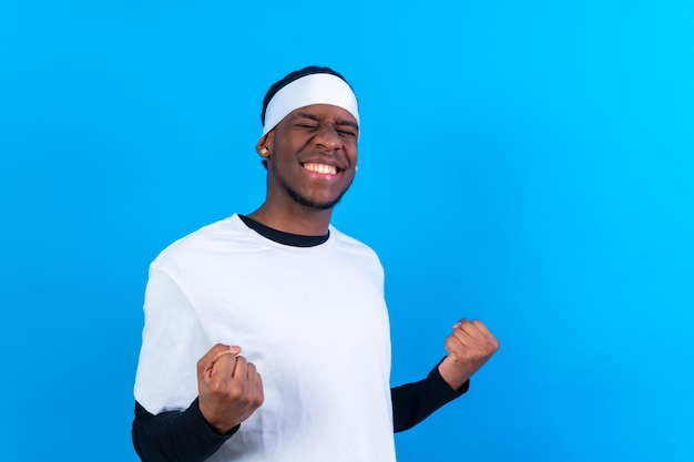 Hombre de etnia negra con ropa blanca de fondo azul haciendo el gesto de victoria y sonriendo