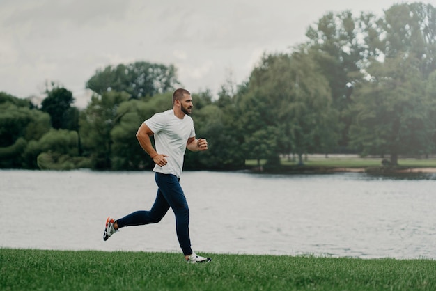 Hombre europeo deportivo vestido con camiseta, pantalones y zapatillas y corre a lo largo del río, entrena al aire libre, lleva un estilo de vida activo, respira aire fresco. Concepto de personas, deporte, bienestar y recreación.