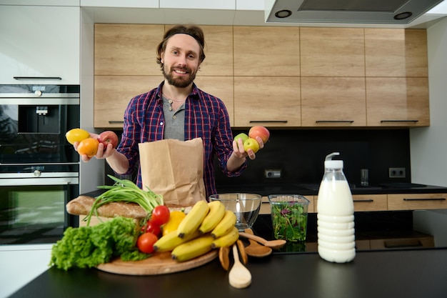 Un hombre europeo guapo y feliz con pantalones a cuadros sostiene frutas frescas crudas en las manos, sonríe mirando a la cámara mientras está parado en la encimera de la cocina detrás de una bolsa de compras ecológica con comida vegana saludable comprada