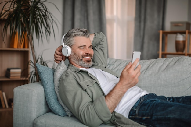 Hombre europeo maduro feliz en auriculares ver video en el teléfono inteligente se encuentra en el sofá y descansa en la vida
