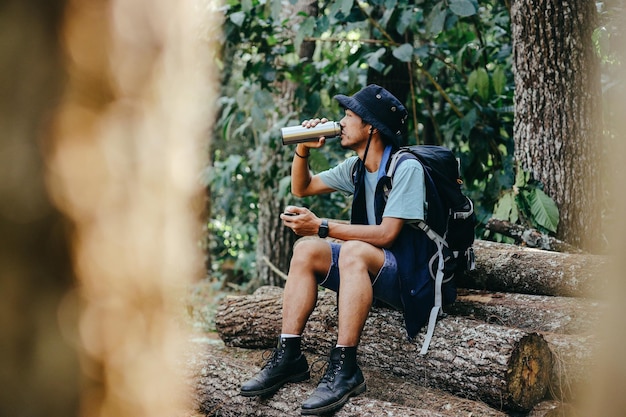 Hombre excursionista asiático cansado sentado en una pila de madera bebiendo agua de una botella mientras descansa durante