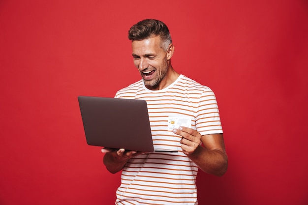 Foto hombre exitoso en camiseta a rayas sonriendo mientras sostiene la tarjeta de crédito y el portátil aislado en rojo