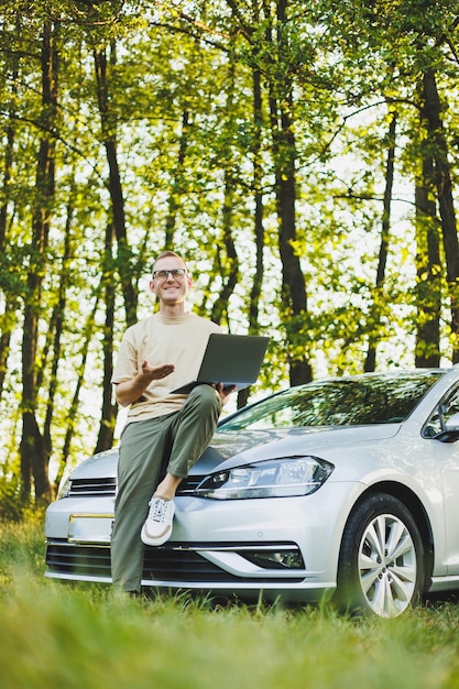 Un hombre exitoso con gafas trabaja en una computadora portátil mientras se apoya en el capó de su coche Trabajo remoto en la naturaleza Trabajar en una laptop en línea Trabajar en la computadora mientras viaja