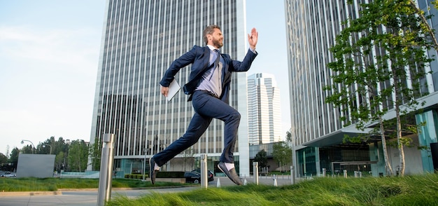 Hombre exitoso en ropa formal saltando con computadora fuera de la oficina, tecnología.