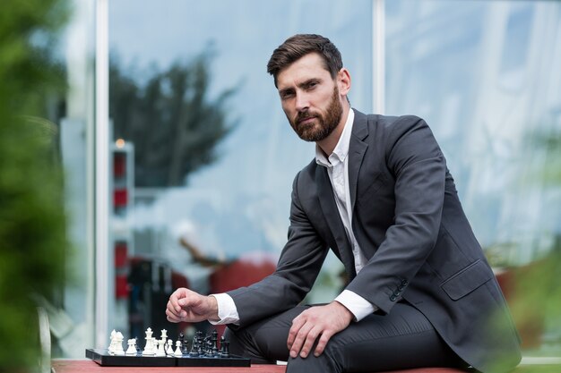 Hombre exitoso sentado en un banco en un traje de negocios jugando al ajedrez y mirando pensativamente a la cámara