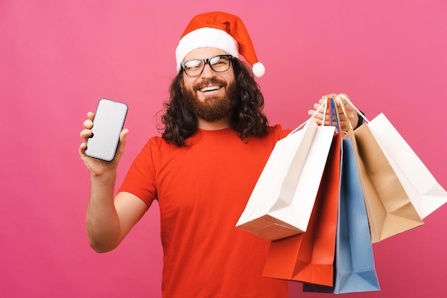 Hombre extasiado con sombrero de Navidad muestra una pantalla de teléfono mientras sostiene bolsas de compras