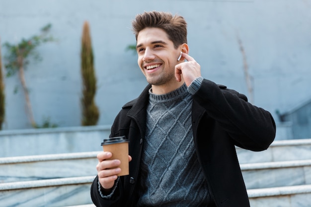 Hombre feliz de 30 años usando audífonos sonriendo y bebiendo café para llevar mientras está sentado en las escaleras de la ciudad