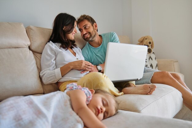 Hombre feliz acariciando el estómago de una mujer embarazada con netbook y sentado en el sofá cerca de la niña durmiendo en la sala de estar en casa