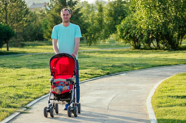 Hombre feliz activo caminando en el parque de verano con un cochecito rojo con su hija