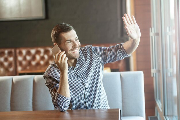 Hombre feliz agitando la mano y hablando por teléfono inteligente en el café