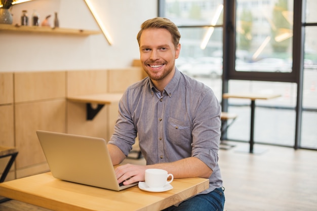 Hombre feliz agradable positivo sentado en la computadora portátil y sonriendo mientras toma un café en la cafetería