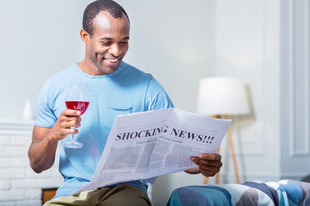 Hombre feliz alegre positivo sonriendo y bebiendo vino mientras lee un periódico