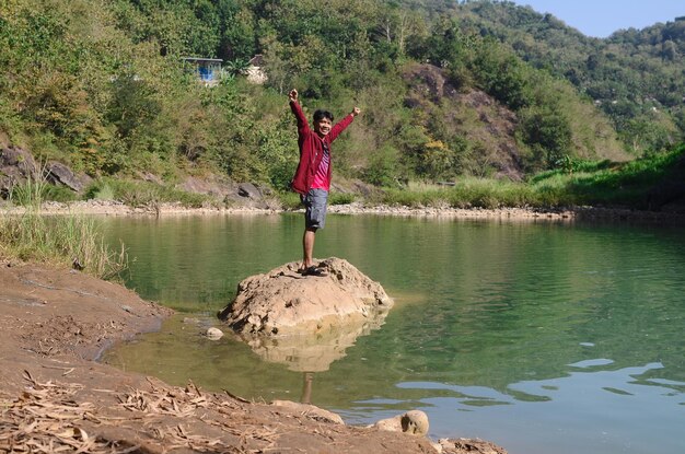 Hombre feliz asiático de pie con chaqueta roja disfrutando de la naturaleza con hermoso río de agua verde
