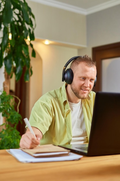 Hombre feliz con auriculares inalámbricos mirando seminarios web, clases de entrenamiento en computadora portátil, tomando notas en un cuaderno de papel en casa