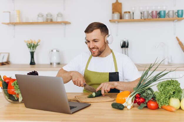 Hombre feliz en auriculares preparando ensalada en la cocina con verduras frescas y mirando la computadora portátil