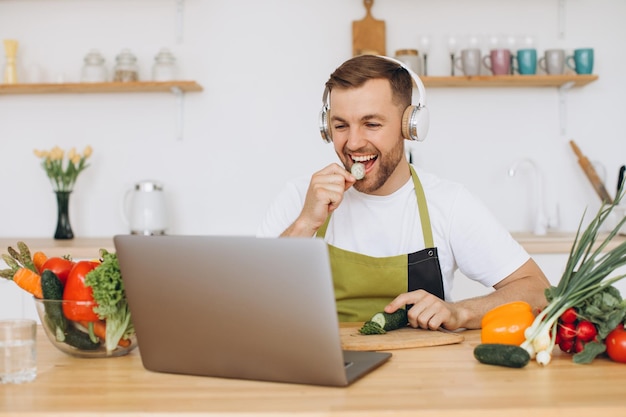 Hombre feliz en auriculares preparando ensalada en la cocina con verduras frescas y mirando la computadora portátil